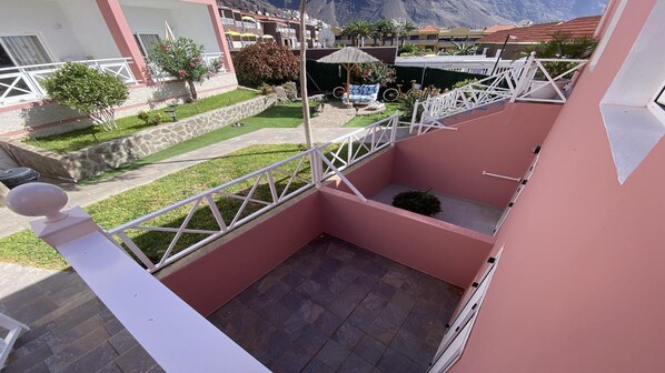 Un balcón con suelo de piedra, una pequeña zona ajardinada con sombrilla de paja y vistas a una cadena montañosa.
