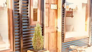 Una puerta de madera con una placa de número, una pequeña planta en maceta y una ventana con persianas horizontales.