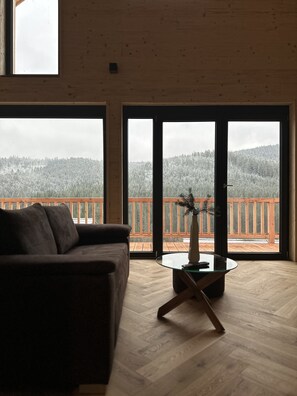 A spacious living room with a large window, a wooden floor, and a brown sofa. - Ferienhaus am Sommerberg (Vöhrenbach)