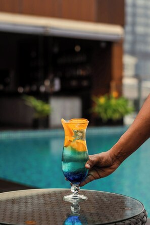 A hand holding a cocktail glass with a layered drink, placed on a table by a poolside.