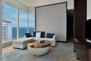 A modern living room with a sofa, a coffee table, a flat-screen TV, and a large window offering a view of the ocean.
