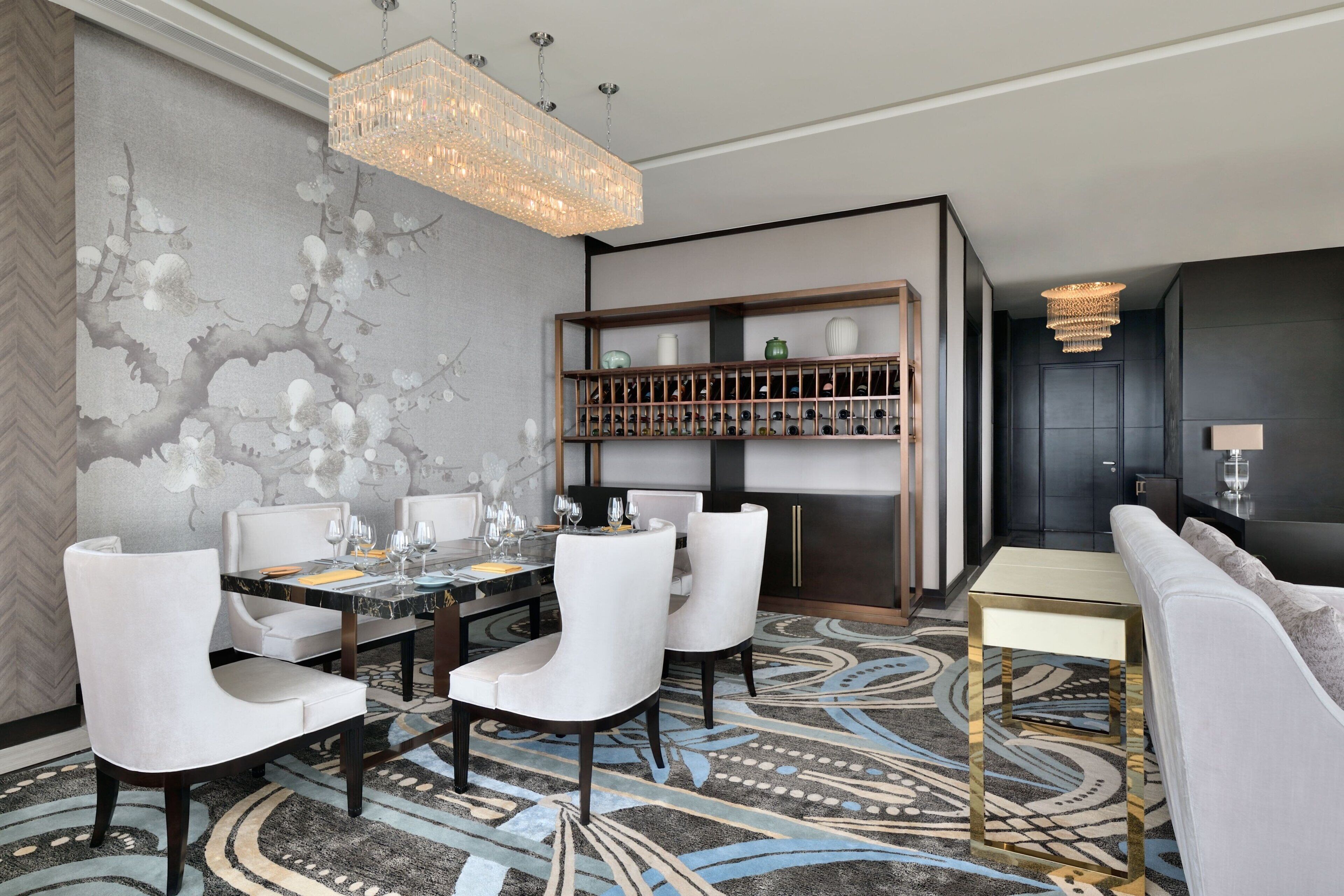 A dining area with a chandelier, a wall-mounted shelf with decorative items, and a patterned rug.
