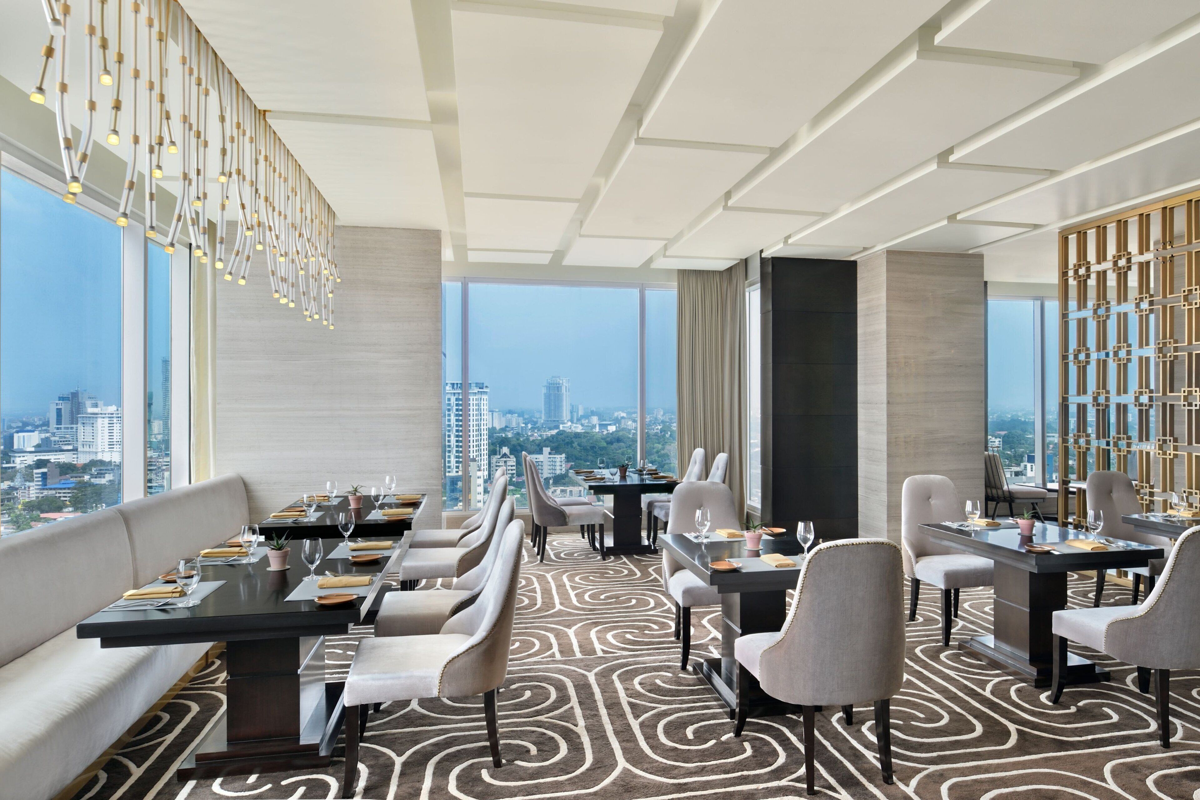 A modern dining area with a city view, featuring a long table with chairs, a patterned carpet, and a decorative ceiling light fixture.