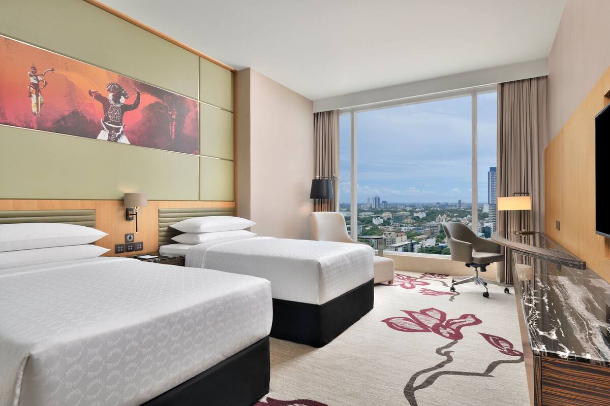 Habitación de hotel con dos camas, un escritorio, una silla, un ventanal con vista a la ciudad y un cuadro en la pared.