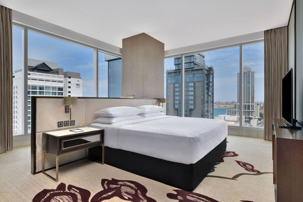 Habitación de hotel moderna con una cama grande, un escritorio y un televisor. Ofrece vistas de la ciudad a través de amplias ventanas.