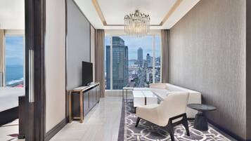 A modern hotel room with a large window offering a city view, a chandelier, a sofa, a TV, and a small table.