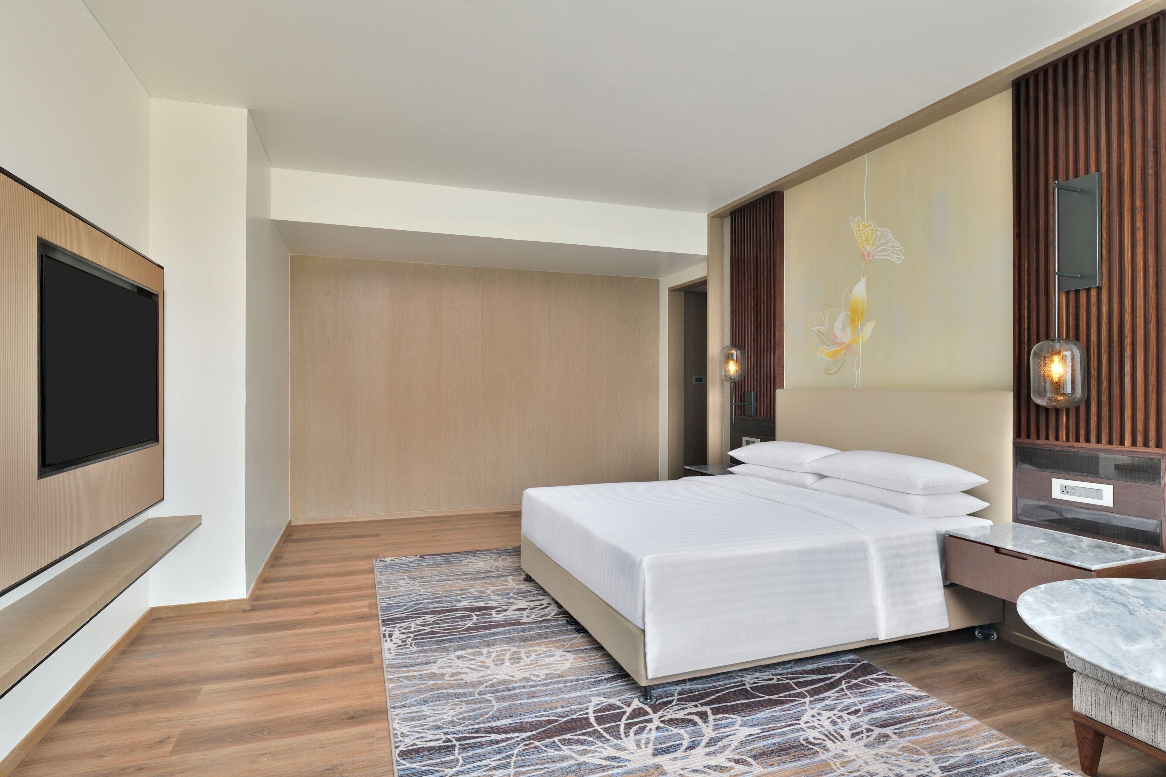 A modern hotel room with a large bed, a flat-screen TV, a marble side table, and a decorative wall panel.