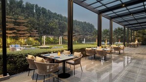 A spacious outdoor dining area with tables and chairs, surrounded by glass panels and a view of a lush garden and hills. - Courtyard by Marriott Gorakhpur (Gorakhpur)