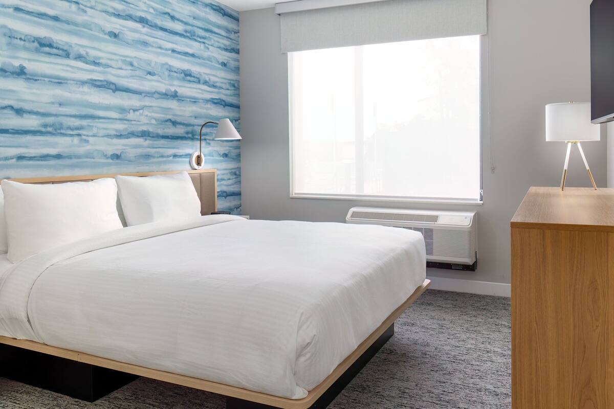 A hotel room with a bed, a nightstand, a lamp, a window with a view, and a wall with a blue pattern.