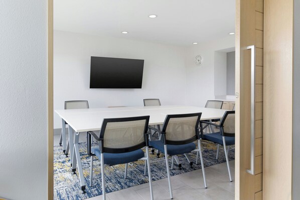 Un salón de conferencias con una mesa blanca, seis sillas, un televisor de pantalla plana montado y un reloj en la pared.