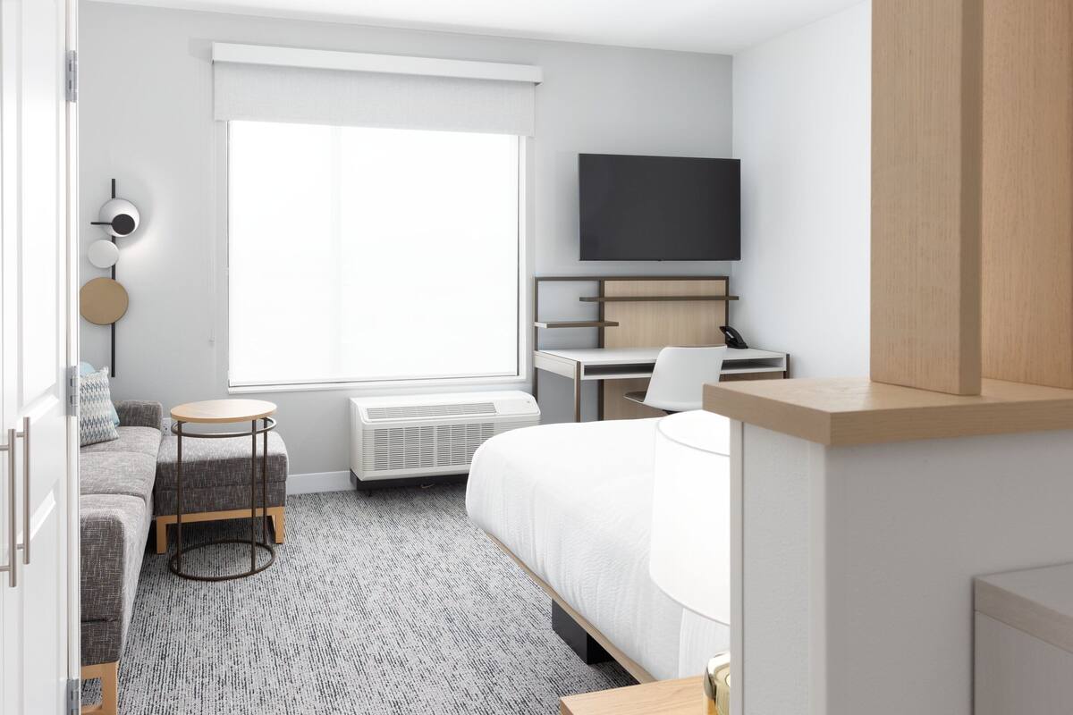 A modern hotel room with a bed, a desk, a chair, a TV, and a round table.