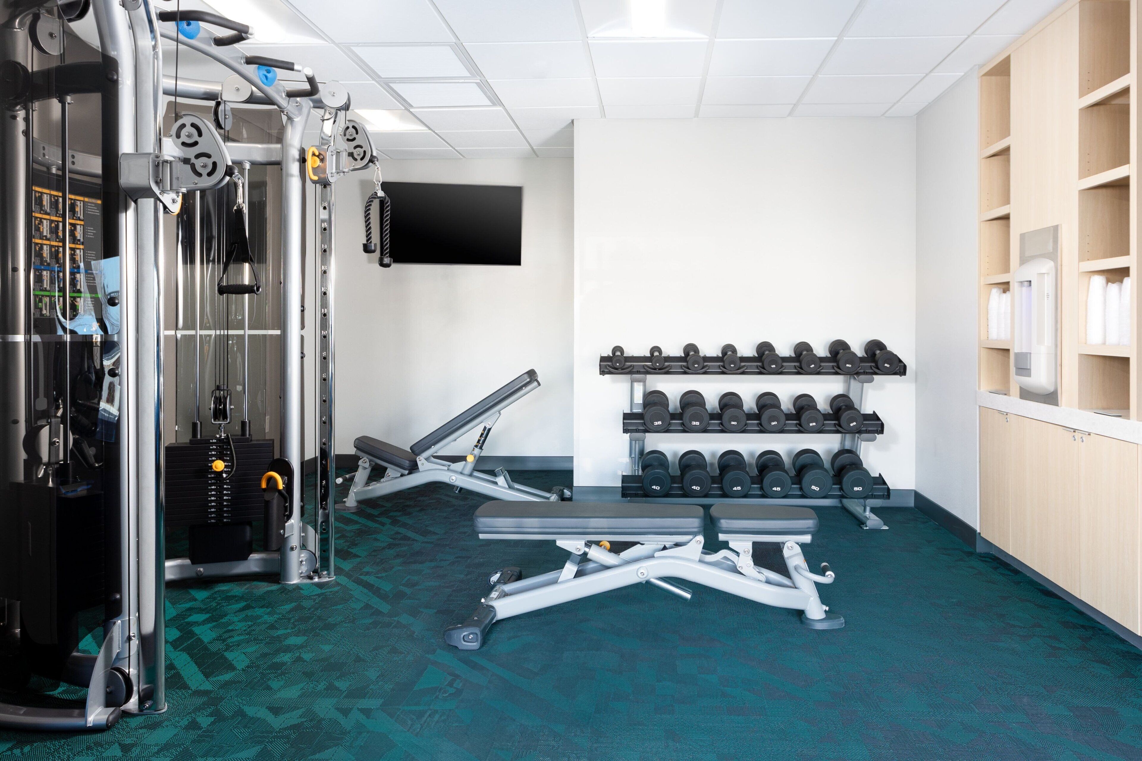 A gym with weightlifting equipment, dumbbells, and a television on the wall.