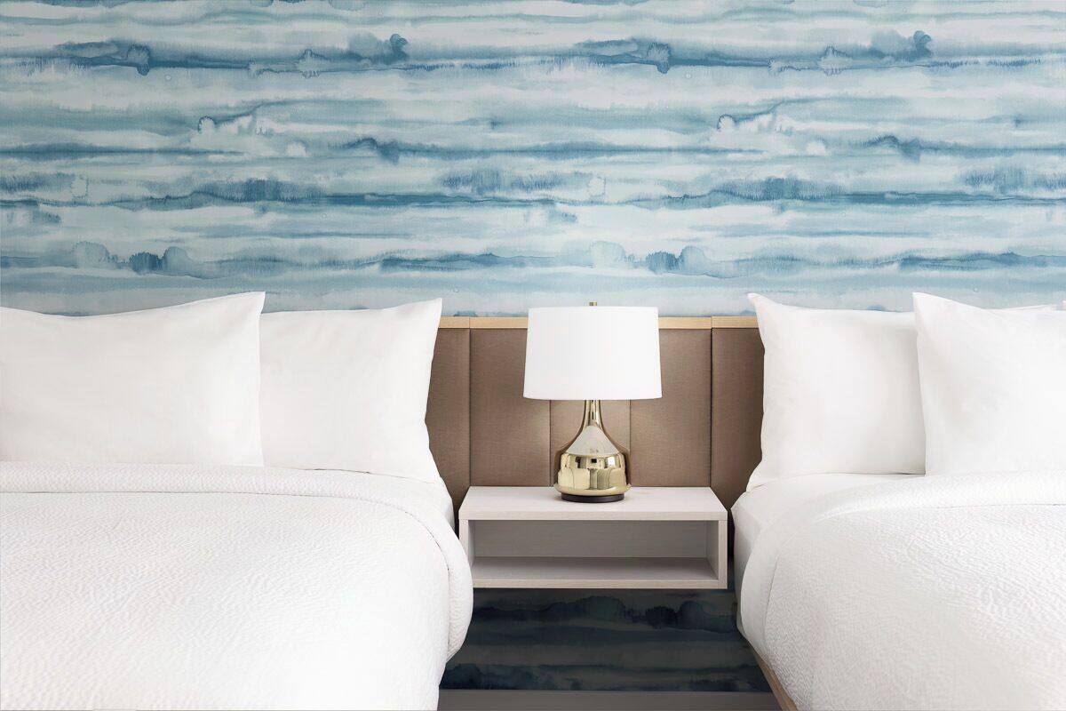 A hotel room with two beds, a nightstand with a lamp, and a wall with a blue abstract pattern.