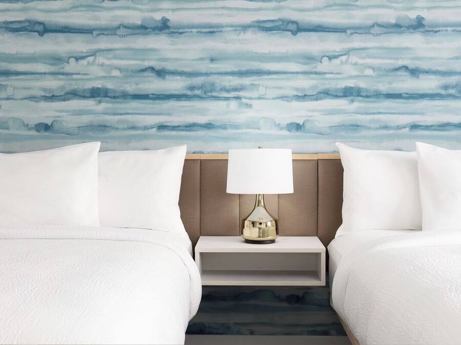 Camera d'albergo con due letti, un comodino con una lampada e una parete decorata con un motivo astratto blu.