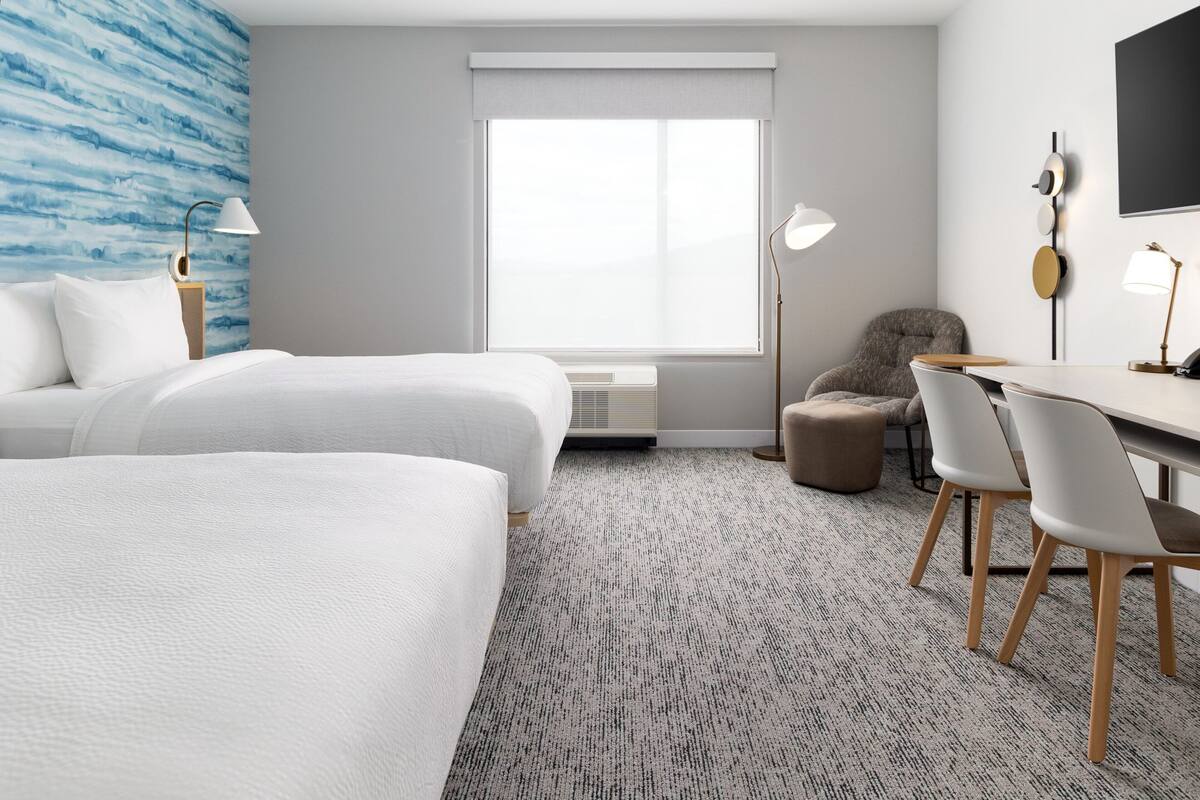 A hotel room with two beds, a desk with chairs, a small armchair, a large window with blinds, and a wall-mounted TV.