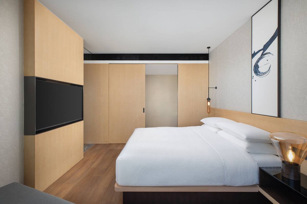 A modern hotel room with a bed, a flat-screen TV, wooden walls, and a large abstract painting.