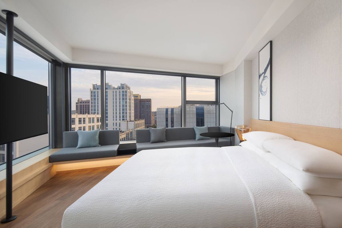 A modern hotel room with a large bed, a seating area with cushions, a flat-screen TV, and a city view through large windows.