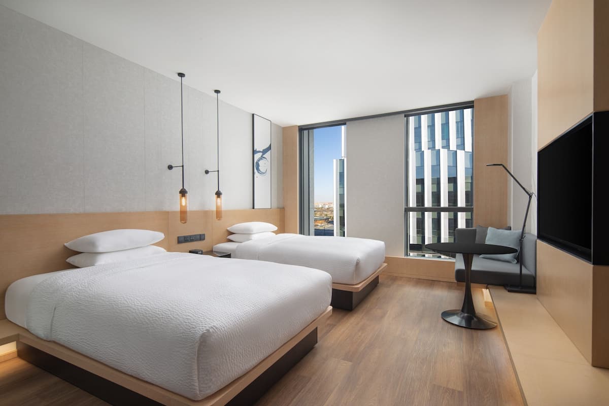 A modern hotel room with two beds, a small table, a sofa, and a large window offering a city view.