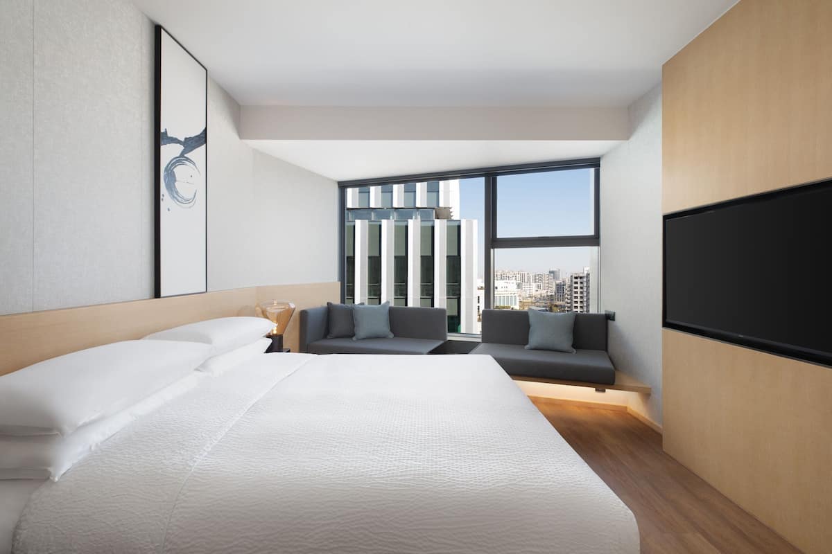 A modern hotel room with a large bed, a sofa, a TV, and a city view.