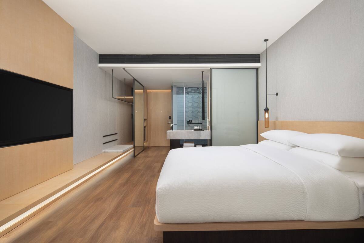 A modern hotel room with a large bed, a built-in TV, a bathroom with a glass shower, and a wooden floor.