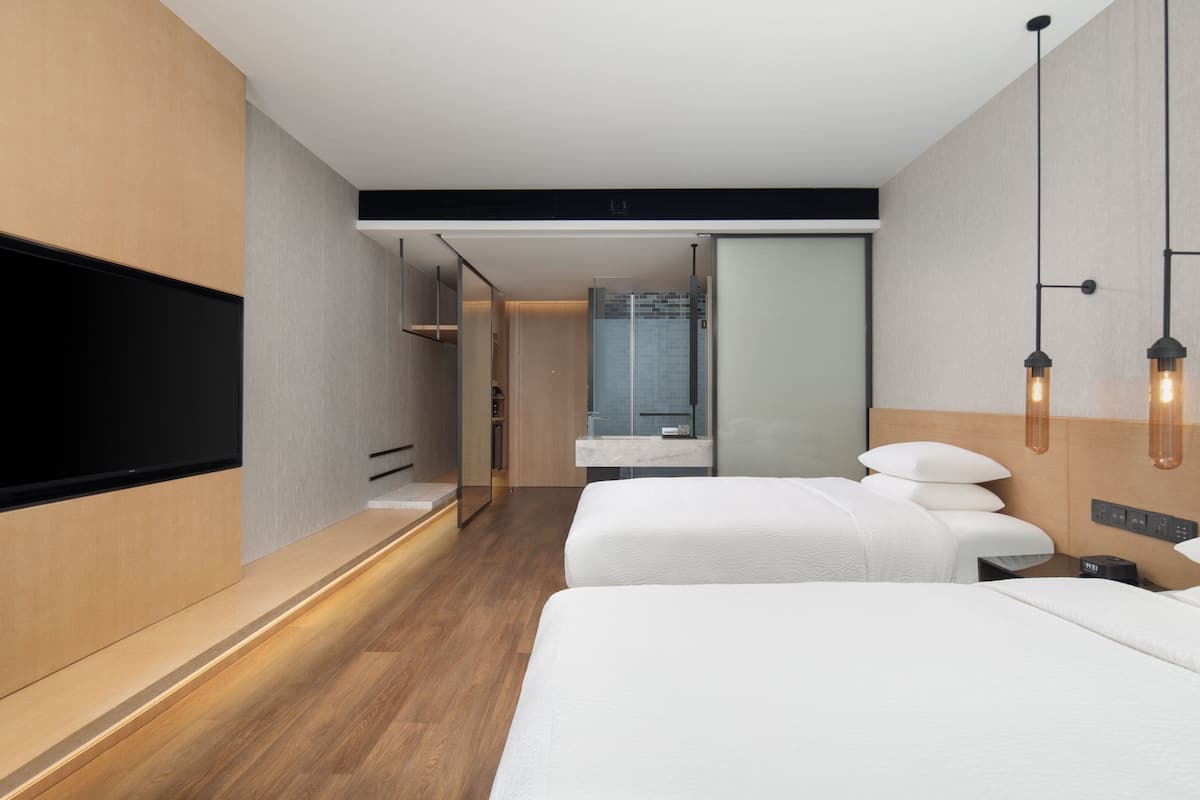 A modern hotel room with two beds, a large flat-screen TV, a wooden floor, and a bathroom with a glass shower.