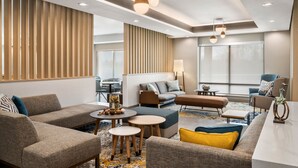 Een moderne hotel lobby met diverse zithoeken, waaronder banken en fauteuils, een salontafel en een kleine eettafel. Op de salontafel staan decoratieve elementen zoals een vaas en een beeldhouwwerk. De ruimte heeft een behang met een patroon en een groot raam dat zorgt voor daglicht.