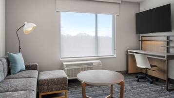 A modern hotel room with a gray sofa, a round table, a desk with a chair, a flat-screen TV, and a large window with a view of trees.