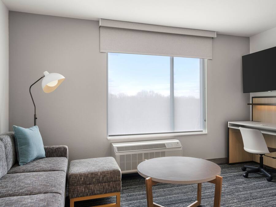 A modern hotel room with a gray sofa, a round table, a desk with a chair, a flat-screen TV, and a large window with a view of trees.