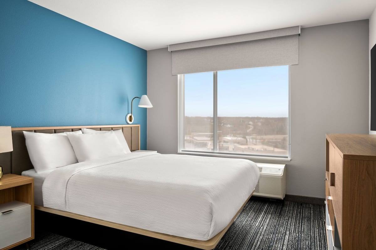 Une chambre d’hôtel avec un grand lit, une table de chevet, une lampe, une fenêtre donnant sur un beau paysage et une commode en bois.