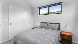 Quarto com uma cama grande, janela com vista para o verde, porta branca e mesa de cabeceira com abajur.