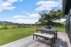 Uma área de refeições ao ar livre com mesa e bancos, cercada por um deck de madeira e vista para um vinhedo.