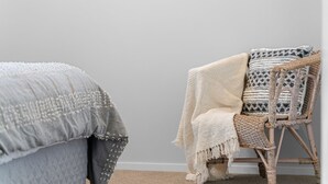 Quarto com cama coberta por um cobertor texturizado, uma cadeira de vime com almofada estampada e uma parede lisa.