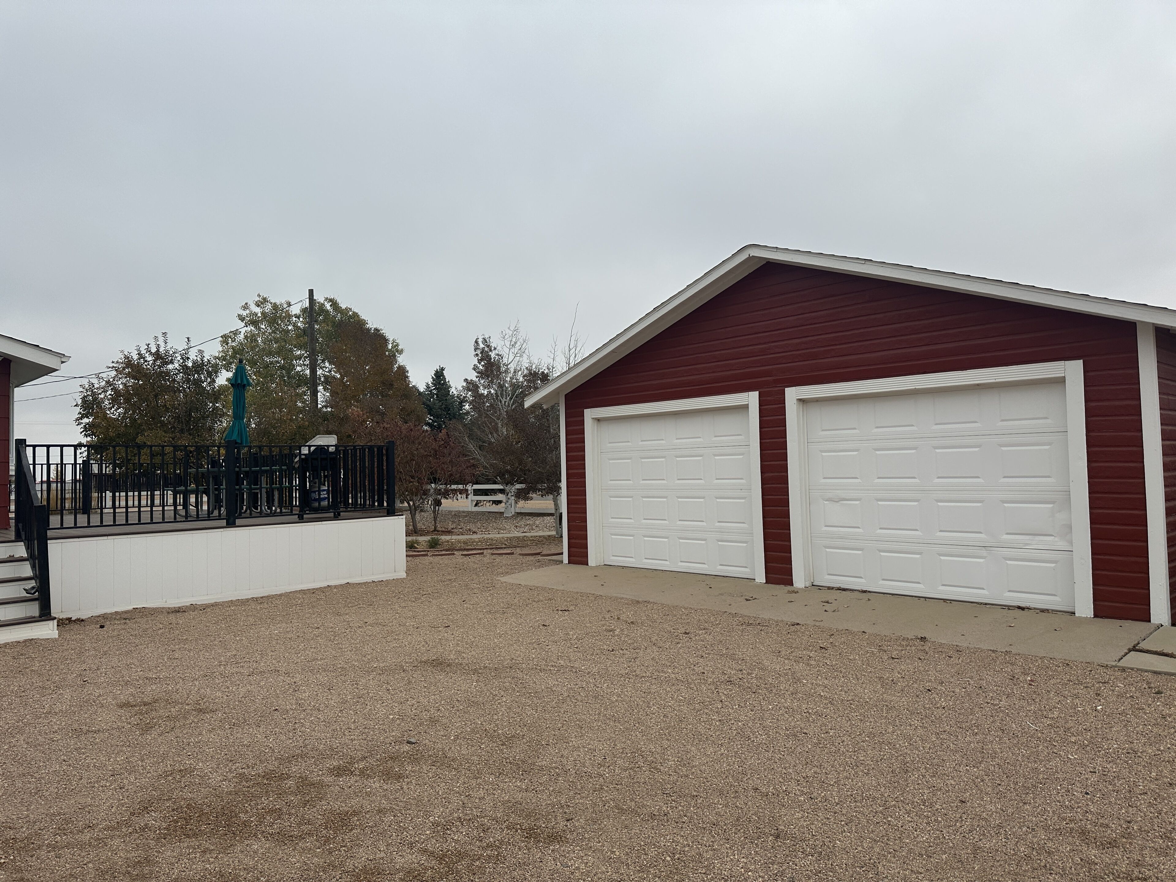 A red building with two white garage doors, a gravel driveway, and a black fence with a green umbrella.