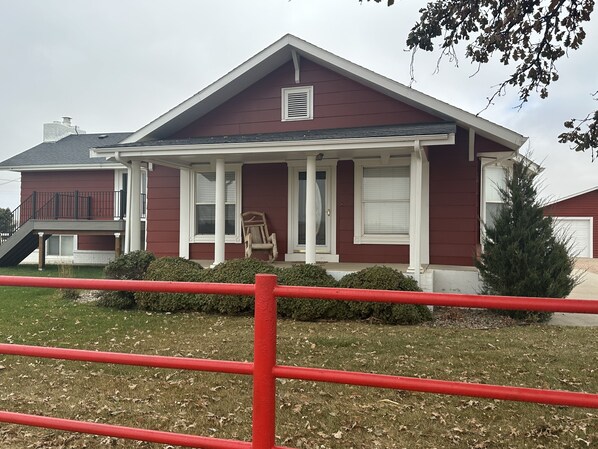 A two-story house with a red exterior and white trim, a front porch, and a small front yard with a red fence. - Adorable Farmhouse within steps of Double Check Arena! (Eaton)