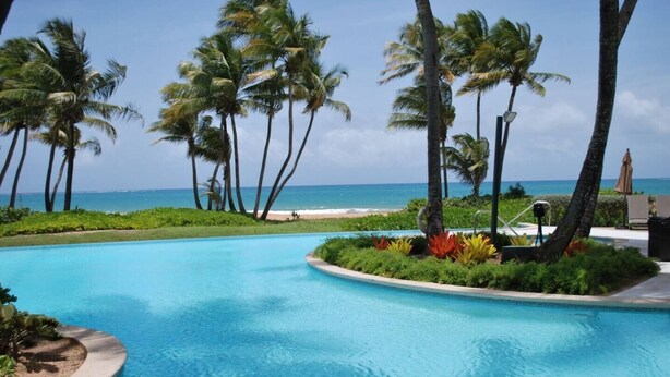 A pool with a curved edge, surrounded by palm trees and a view of the beach. - Resort Condo at Wyndham Grand Rio Mar (Río Grande)