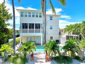 A two-story building with a pool, surrounded by palm trees and a white fence. - NEW! The Oasis on Primo! Next To Margaritaville & In The Heart of Time Square! (Fort Myers Beach)