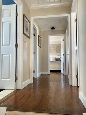 A hallway with wooden flooring leading to a bedroom with a bed and a nightstand. - NEW! The Oasis on Primo! Next To Margaritaville & In The Heart of Time Square! (Fort Myers Beach)