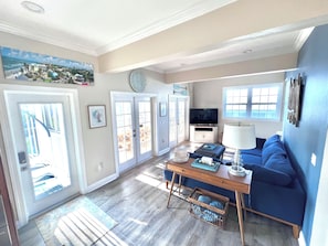 A living room with a blue sofa, a wooden coffee table, a television, and a large window. - NEW! The Oasis on Primo! Next To Margaritaville & In The Heart of Time Square! (Fort Myers Beach)