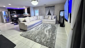 A modern living room with a sectional sofa, a flat-screen TV, and a kitchen area in the background.