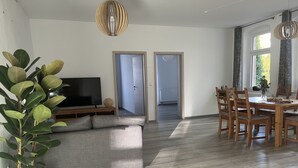 Ein modernes Wohnzimmer mit grauem Sofa, einem Flachbildfernseher, einem Esstisch mit Holzstühlen und einer Topfpflanze.