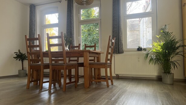 Ein Essbereich mit Holztisch und -stühlen, großen Fenstern mit Vorhängen, einem Heizkörper und Topfpflanzen.