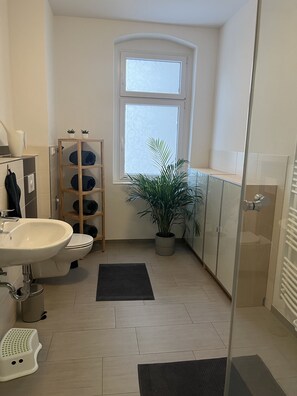Ein modernes Badezimmer mit Waschbecken, Toilette und Duschbereich. Ein Fenster bietet Blick ins Freie, ein Regal mit zusammengerollten Handtüchern und ein Topf mit einer Pflanze sind ebenfalls vorhanden.