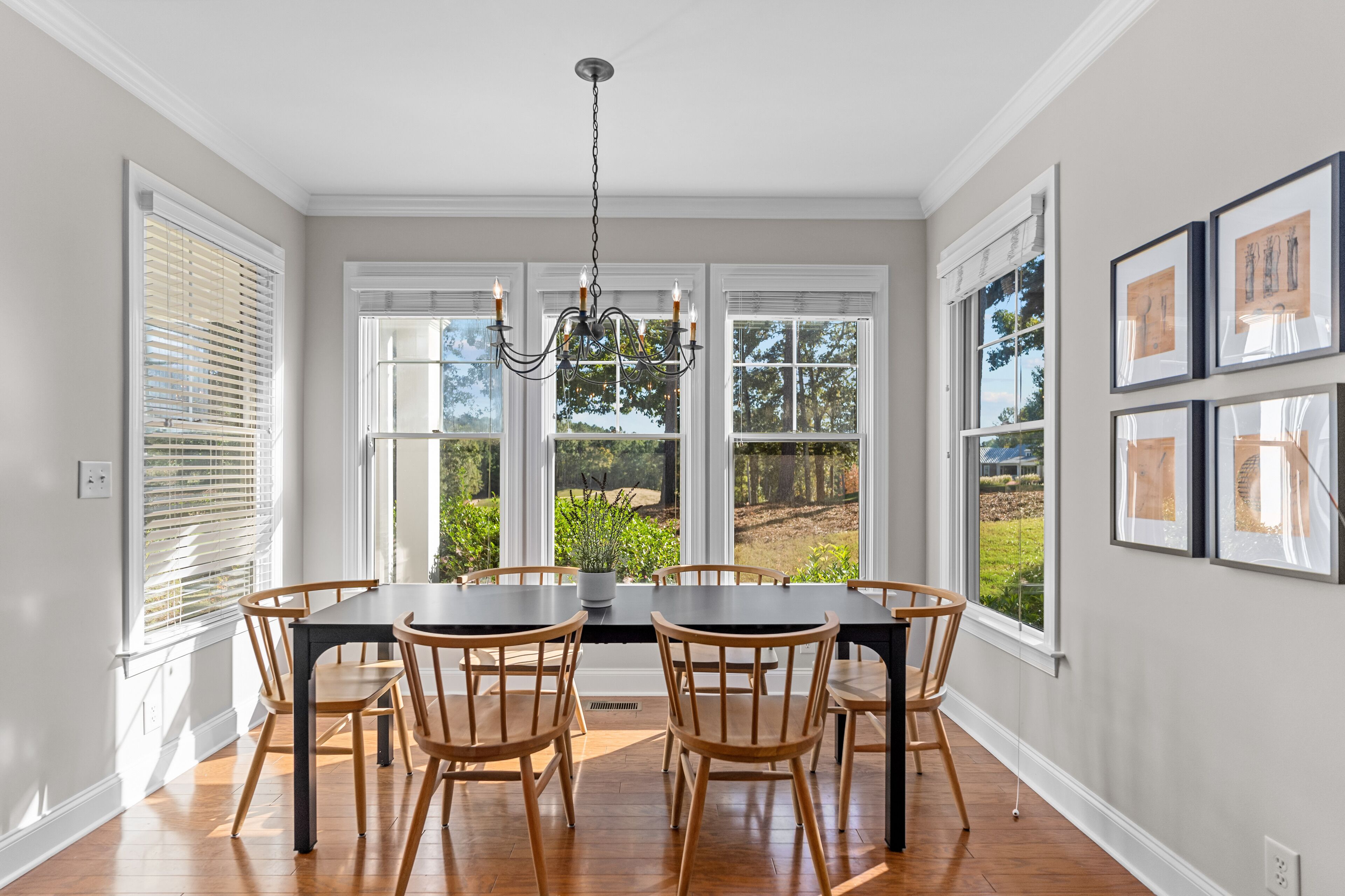 A dining room with a large table, six chairs, a chandelier, and large windows with blinds.