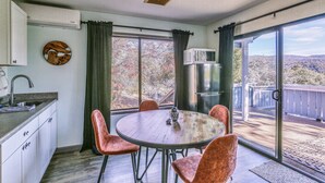 Un comedor con una mesa redonda y cuatro sillas, una cocina con un lavamanos y alacenas, y un amplio ventanal corredizo que da a una terraza con vistas a árboles y colinas.