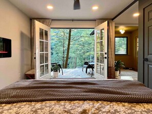 A bedroom with a large bed, a view of a forest through open doors, and a small balcony with chairs and a table.