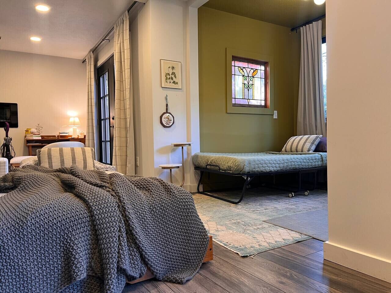 A cozy bedroom with a bed, a smaller bed, a window with curtains, a wall-mounted lamp, and a framed picture.