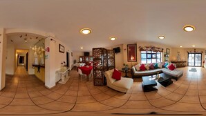 Un espacioso vestíbulo de hotel con techo curvo, piso de baldosas, mostrador de recepción, zona de estar con sofás y sillones, y elementos decorativos como mariposas y cuadros en la pared.