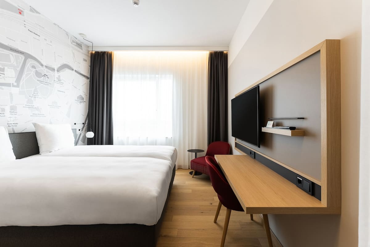 A hotel room with a large bed, a desk with a TV, a red chair, and a map on the wall.