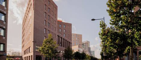 A modern urban street with tall brick buildings, a clear sky, and green trees lining the sidewalk.