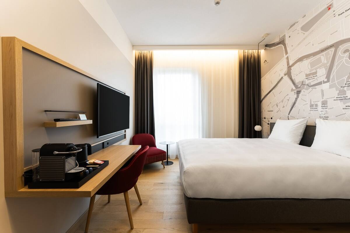 A hotel room with a bed, a desk with a television, a red chair, and a map on the wall.